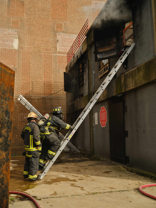 Feuerwehrleute in Helmen, die eine Leiter zu einem raucherfüllten Gebäude hinaufsteigen, mit einem Metallobjekt links und Rohren auf dem Boden.