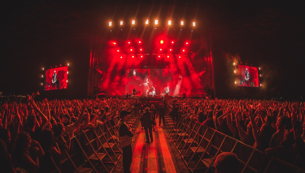 Eine große Menschenmenge steht vor einer hell erleuchteten Bühne mit Geländern und schaut Musikern zu, während andere die Szene beobachten.