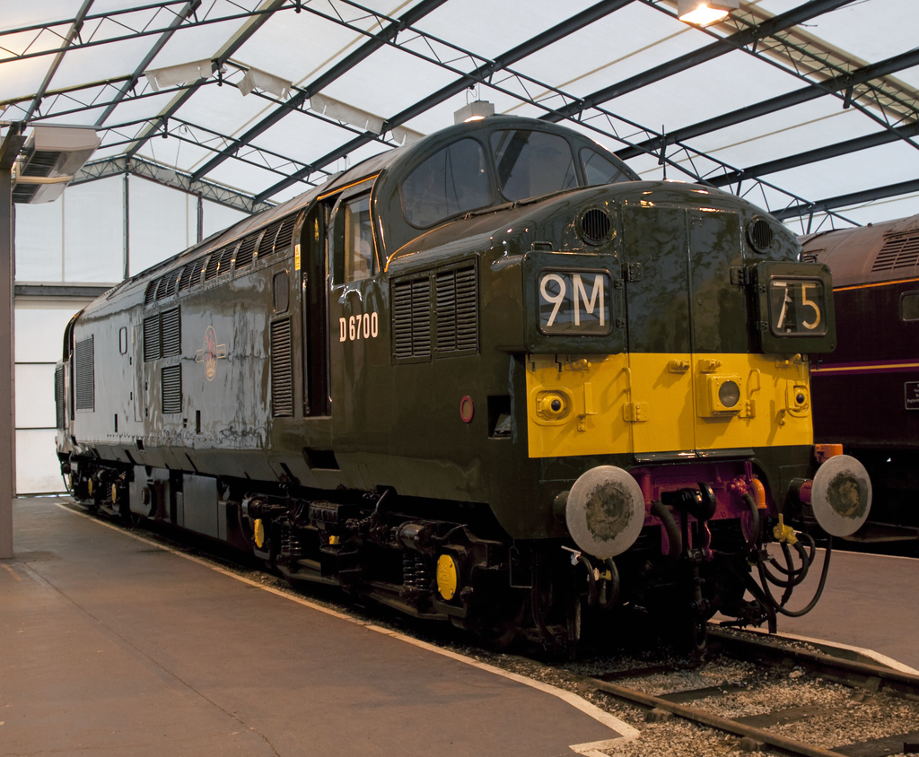 Ein Zug fährt auf den Schienen mit einem anderen Zug auf der rechten Seite neben einem Bahnsteig, an dem ein Mast mit Metallstangen und Lampen befestigt ist.