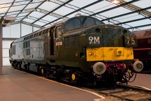 Ein Zug fährt auf den Schienen mit einem anderen Zug auf der rechten Seite neben einem Bahnsteig, an dem ein Mast mit Metallstangen und Lampen befestigt ist.