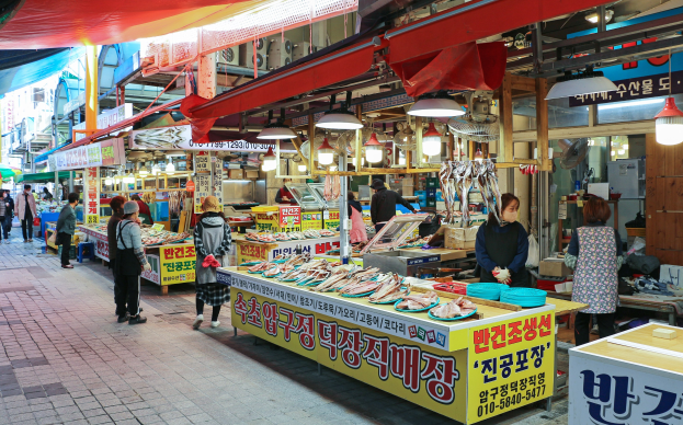 Ein belebter Straßenmarkt in Seoul, Südkorea, mit Menschen, die gehen, Ständen mit verschiedenen Gegenständen und Gebäuden im Hintergrund unter einem klaren blauen Himmel.