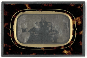 Ein Schwarz-Weiß-Foto eines Mannes in Anzug und Krawatte und einer Frau in unbekannter Kleidung, die ernst vor einem alten Fernseher gegen einen weißen Hintergrund stehen.