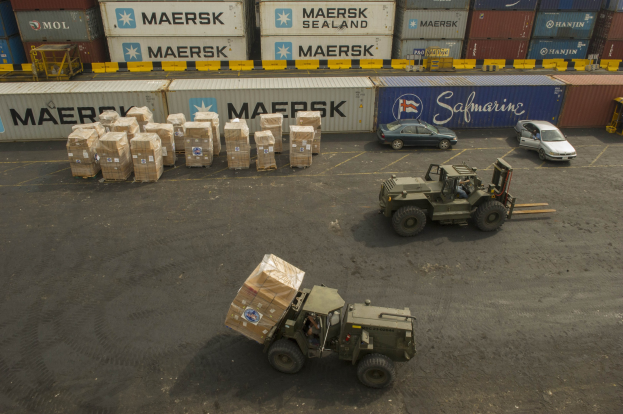 Militärfahrzeug vor einem großen Container Schiff mit mehreren Fahrzeugen und verstreuten Kartonagen auf der Straße und Containern mit Text im Hintergrund.
