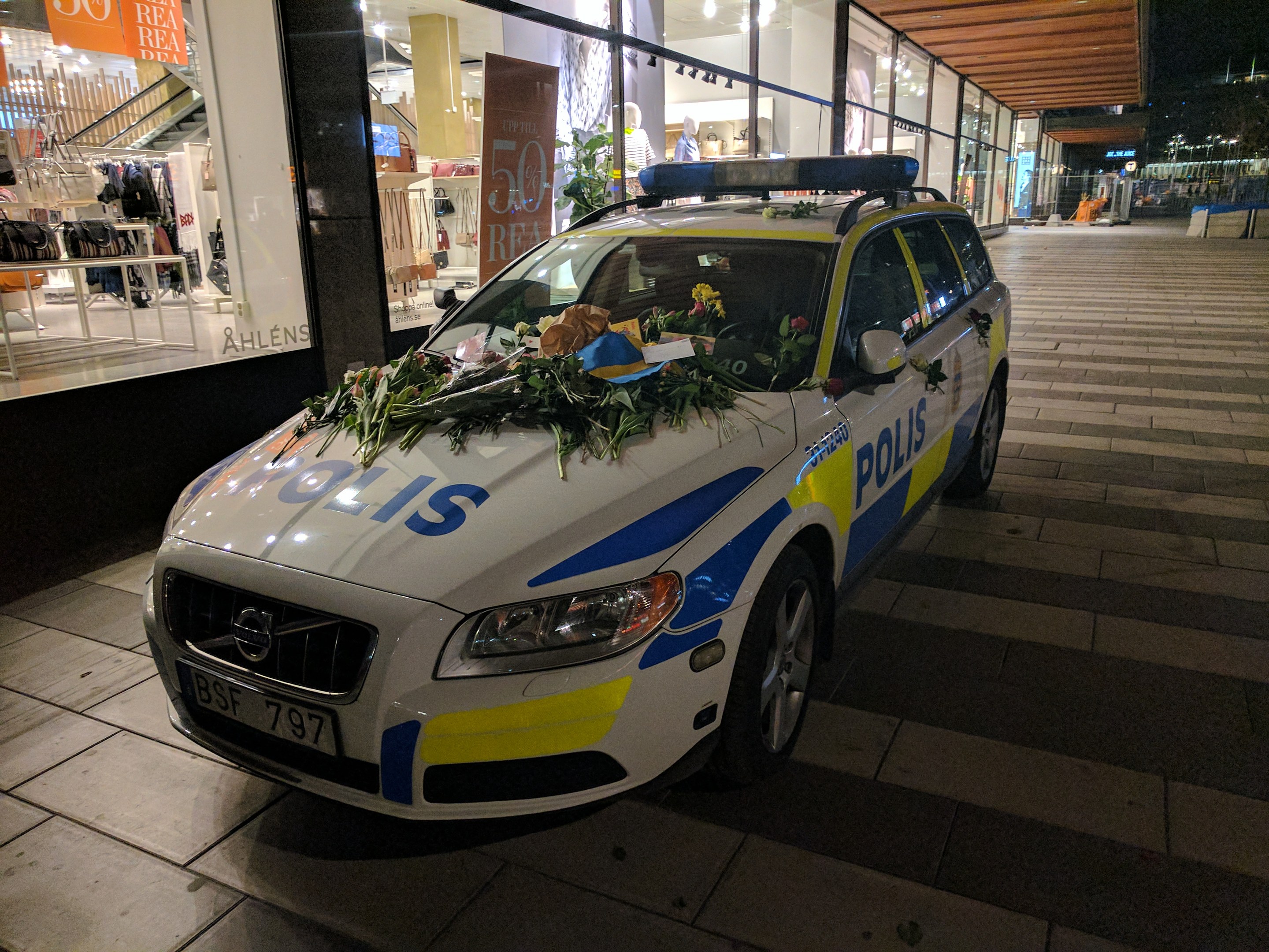 Ein mit Blumen und Blättern geschmückter Polizeiwagen parkt vor einem Geschäft mit Glaswänden bei Nacht und zeigt Schaufensterpuppen in Kleidern und beleuchtete Schilder im Inneren.