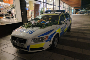 Ein mit Blumen und Blättern geschmückter Polizeiwagen parkt vor einem Geschäft mit Glaswänden bei Nacht und zeigt Schaufensterpuppen in Kleidern und beleuchtete Schilder im Inneren.