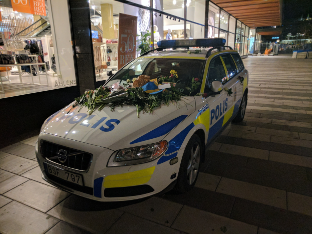 Ein mit Blumen und Blättern geschmückter Polizeiwagen parkt vor einem Geschäft mit Glaswänden bei Nacht und zeigt Schaufensterpuppen in Kleidern und beleuchtete Schilder im Inneren.