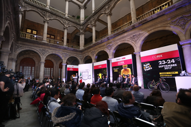 Eine Gruppe von Menschen, die an einem Tisch mit einem Rednerpult und einem Mikrofon vor einer Menge, Kameras und Bannern sitzen, was auf eine Pressekonferenz für das CDMX 2017-Event hindeutet.