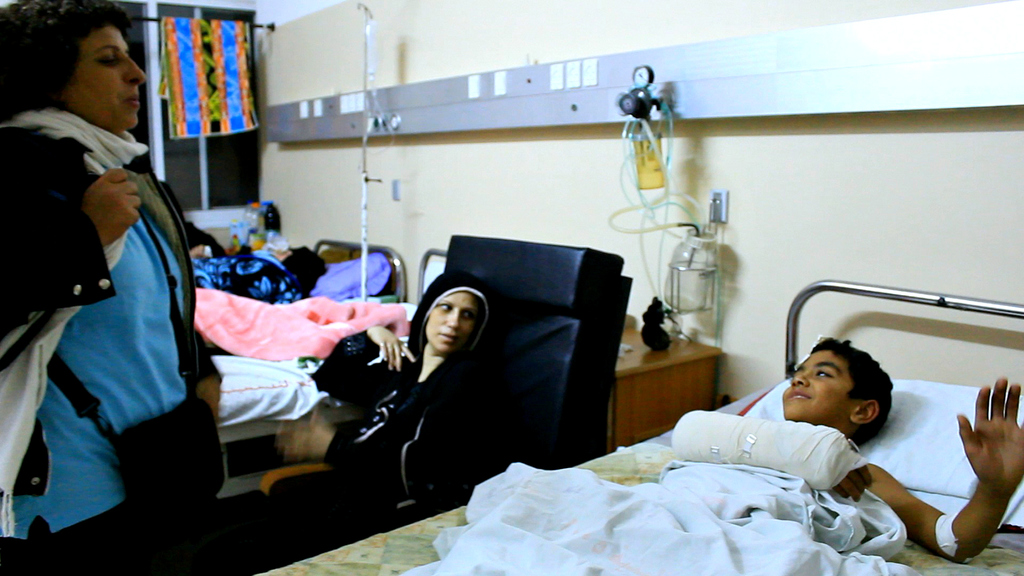 Ein Krankenhauszimmer mit mehreren Betten, von denen jedes von einem Patienten belegt ist, Kochsalzflaschen auf Ständern zwischen den Betten und eine Frau mit einer Tasche in der Nähe eines der Patienten.