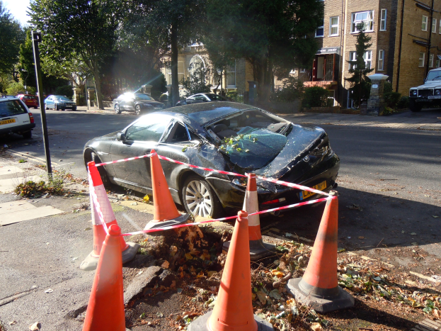 Ein beschädigtwagen am Straßenrand umgeben von orangen Verkehrskegeln, mit Bäumen, Gebäuden und einem klaren blauen Himmel im Hintergrund.
