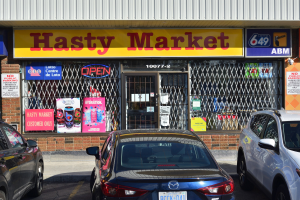 Ein geschäftiges Hasty Market-Geschäft in Toronto, Ontario, mit Autos auf der Straße vor seinen Glaswänden und -türen, die Plakate mit Text zeigen.