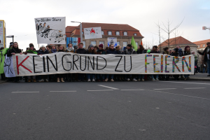 Demonstranten mit einem Transparent mit der Aufschrift 'Kein Grund zur Feier' gegen deutsche Sparmaßnahmen auf einer Stadtstraße mit Gebäuden, Bäumen und einem klaren Himmel.