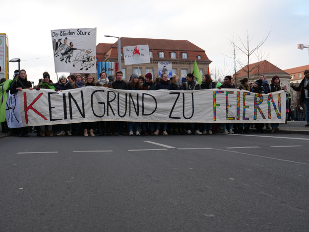 Demonstranten mit einem Transparent mit der Aufschrift 'Kein Grund zur Feier' gegen deutsche Sparmaßnahmen auf einer Stadtstraße mit Gebäuden, Bäumen und einem klaren Himmel.