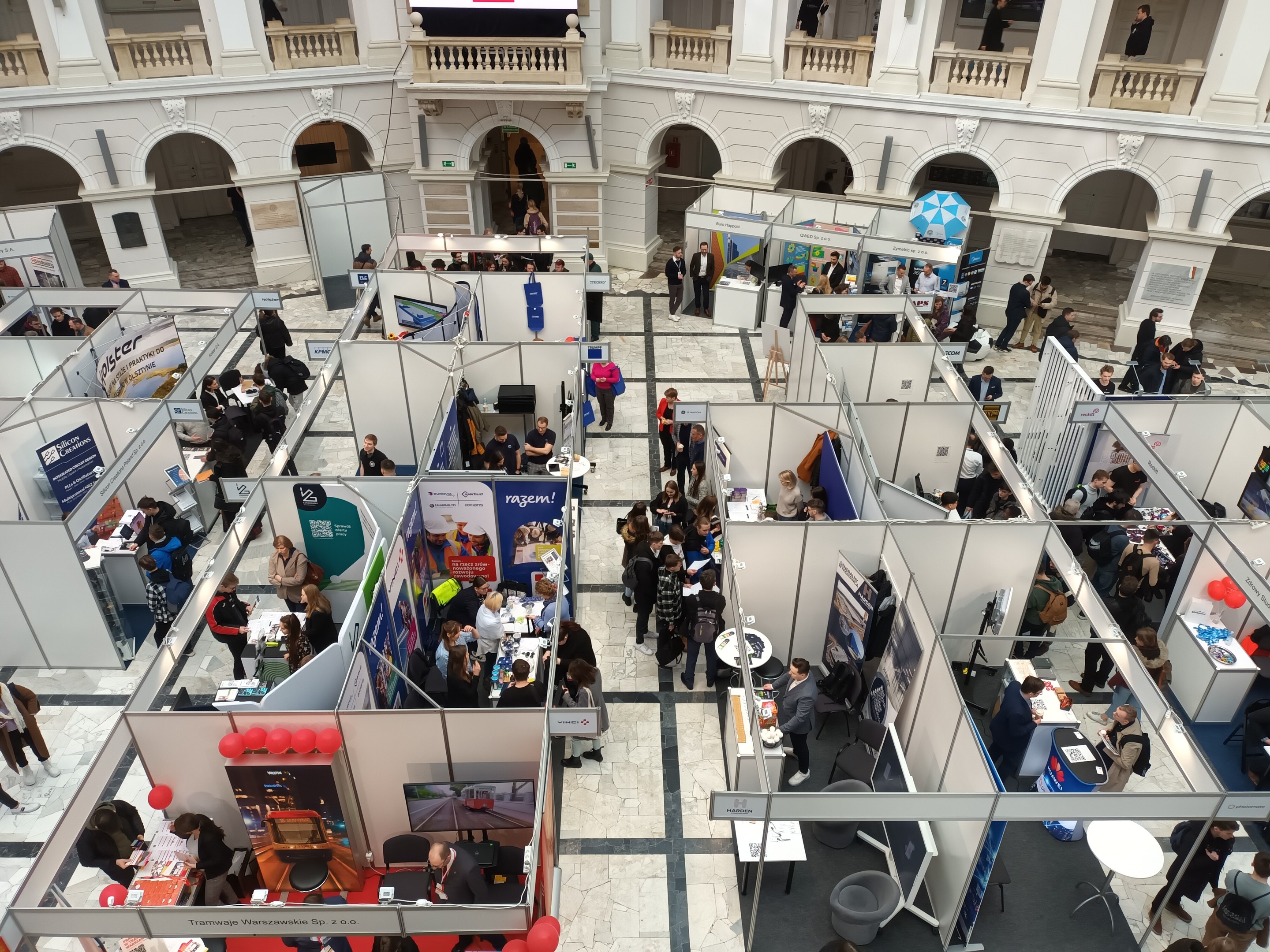 Große Gruppe von Teilnehmern auf einer internationalen Fachmesse mit Ständen, Bannern, Tischen, Stühlen und Ballons in einem Raum mit architektonischen Bögen und Säulen.