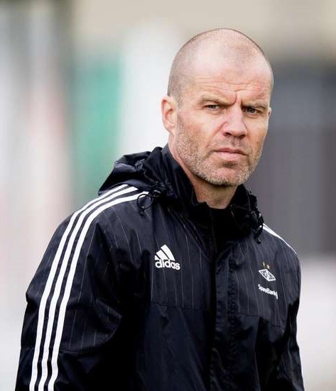 Mann mit Glatze in einem blauen Jackett mit weißen Streifen und weißem Kragen, der auf einem Fußballfeld steht und nachdenklich die Hände zusammenlegt.