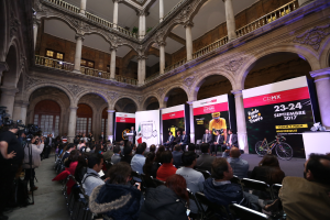 Eine Gruppe von Menschen sitzt an einem Tisch mit einem Podium und einem Mikrofon vor einer Menge, Kameras und Transparenten, was auf eine Pressekonferenz für das CDMX 2017-Event hindeutet.