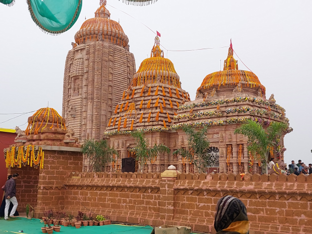 Eine Gruppe von Menschen vor einem farbenfrohen Hindu-Tempel mit Girlanden und Flaggen, die sich auf einem grünen Teppich unter einem hellblauen Himmel mit üppiger Vegetation versammelt haben.
