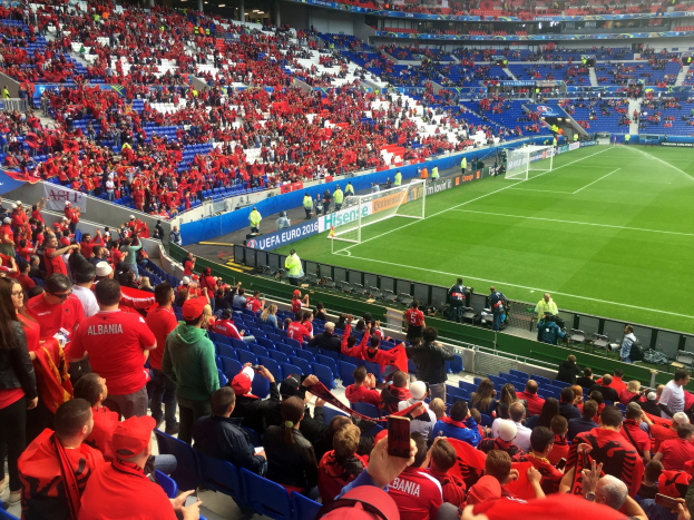 Eine große Menge Menschen in einem Stadion schaut ein Fußballspiel, mit Sitzplätzen und Stehplätzen, Bannern, einem Torpfosten und Deckenbeleuchtung.