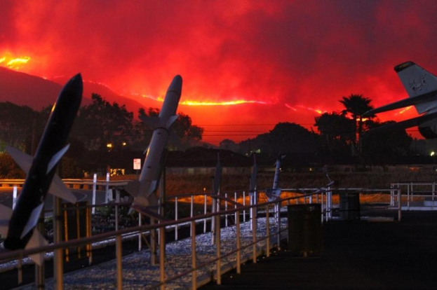 Ein Flugzeug fliegt über eine mit Raketen ausgestattete Start- und Landebahn, umgeben von Zäunen, Mülltonnen, Bäumen, Laternenmasten und Gebäuden, mit einem von den kalifornischen Waldbränden verqualmten Himmel im Hintergrund.