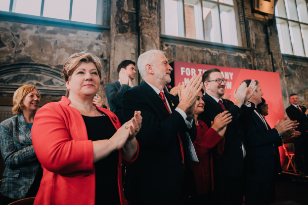 Eine Gruppe von Menschen steht vor einer Menge und klatscht, während im Hintergrund ein Banner zu sehen ist, das likely einen Wahlsieg anzeigt.