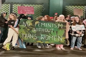 Eine Gruppe von Menschen hölt ein Transparent mit der Aufschrift "Kein Feminismus ohne trans Frauen" und Schilder in einem öffentlichen Raum mit einer Wand und einem Rollladen im Hintergrund.