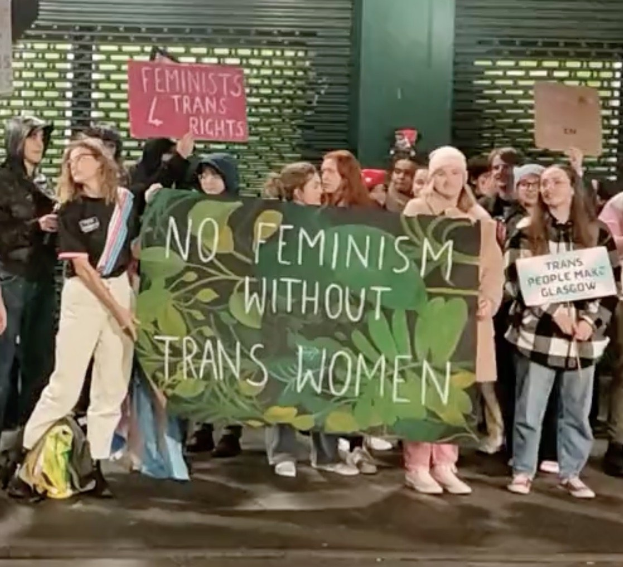 Eine Gruppe von Menschen hölt ein Transparent mit der Aufschrift "Kein Feminismus ohne trans Frauen" und Schilder in einem öffentlichen Raum mit einer Wand und einem Rollladen im Hintergrund.