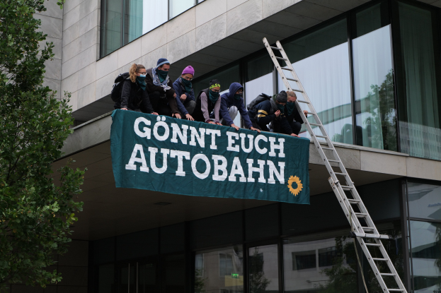 Gruppe von Menschen auf einem Gebäude, die ein Transparent halten, mit einem Baum links und Glasfenstern im Hintergrund, die scheinbar für die deutsche Regierung protestieren.