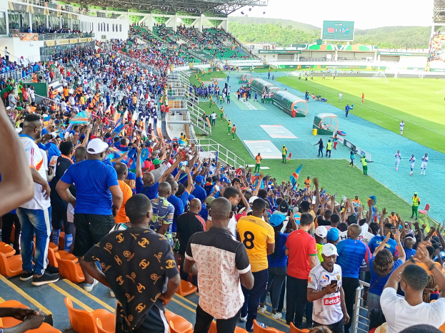 Eine große Menschenmenge versammelt sich vor einem eingezäunten Fußballfeld mit Hügeln und einem klaren blauen Himmel im Hintergrund und erwartet den Beginn eines Spiels während der African Cup of Nations 2019.