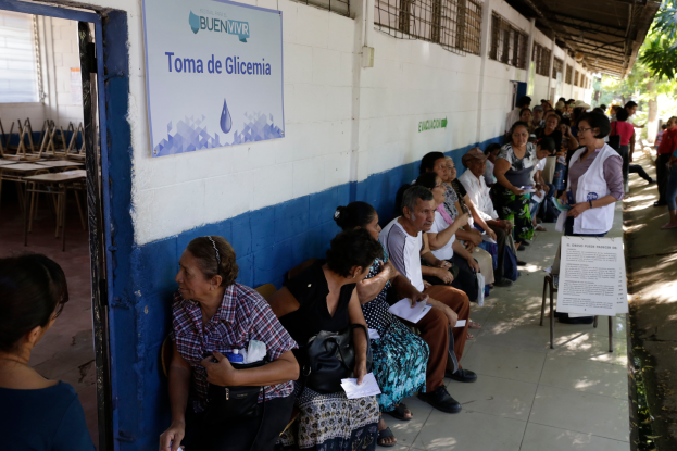 Eine Gruppe von Menschen sitzt auf Bänken vor einem Gebäude und wartet in einer Schlange an einer Wahllokal in Guadalajara, Mexiko, mit einer Texttafel an der Wand und Bäumen im Hintergrund.