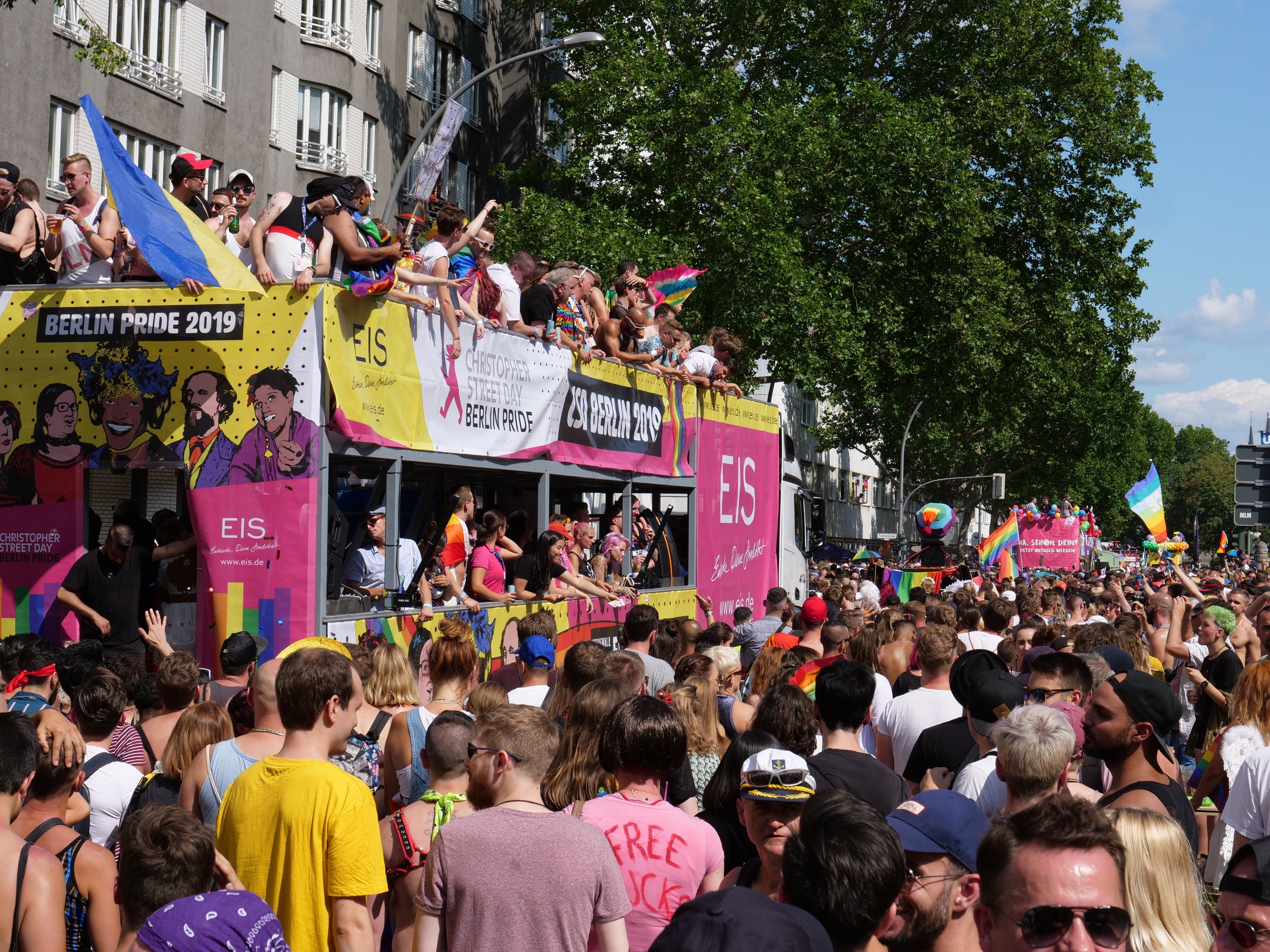 Eine große Menge marschiert während des Berlin Pride 2019 die Straße entlang, viele tragen Kappen und Schutzbrillen, einige halten Fahnen, mit Bannern, Laternenmasten, Bäumen und Gebäuden im Hintergrund unter einem bewölkten Himmel.