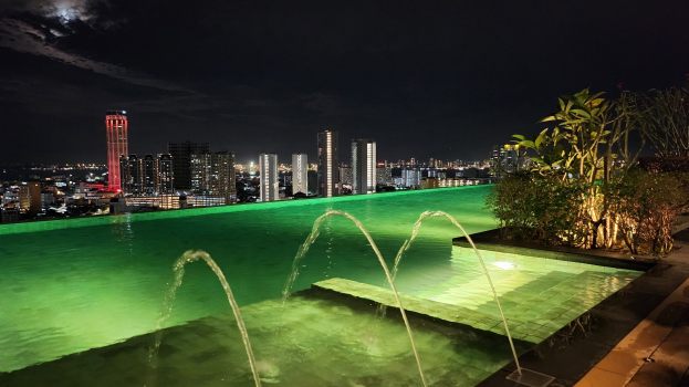 Ein Schwimmbad mit einem zentralen Brunnen, umgeben von Pflanzen auf der rechten Seite und Gebäuden im Hintergrund, beleuchtet von Stadtlichtern in der Nacht.