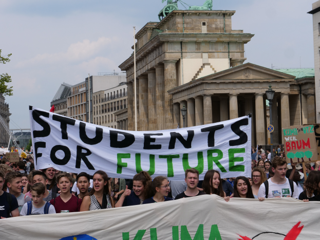 Eine Gruppe von Schülern marschiert in Berlin, eine bunt bemalte Fahne haltend, auf der "Students for Future" steht, mit Gebäuden, Bäumen und Himmel im Hintergrund.