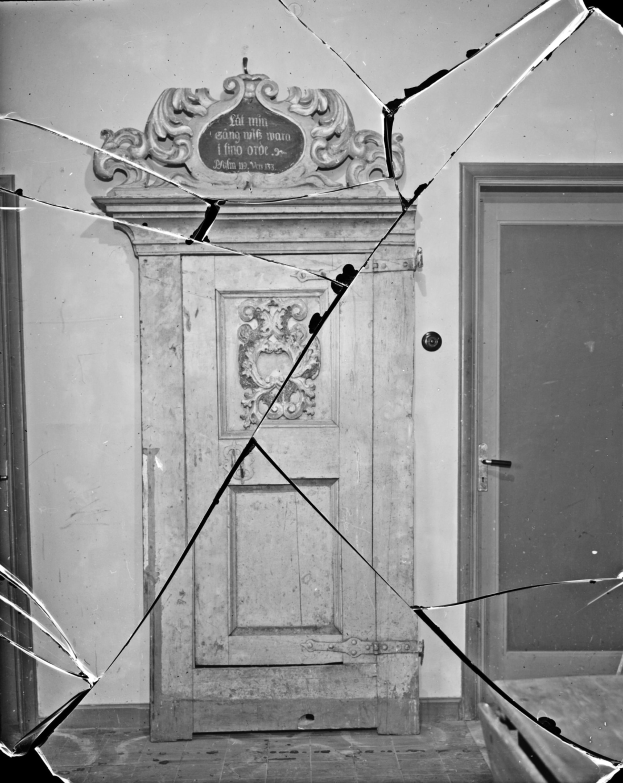 Ein Schwarz-Weiß-Foto einer zerbrochenen Tür in einem Raum mit verstreuten Glasscherben drumherum und einer Wand im Hintergrund.