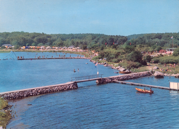 Altes Schwarz-Weiß-Foto eines Sees mit einem Boot, einer Brücke, Flaggen, Felsen, Bäumen, Gebäuden und Menschen im Wasser.