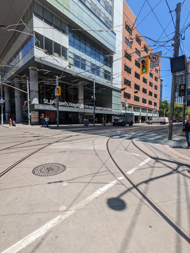 Stadtstraße mit einer durch sie verlaufenden Bahnschiene, umgeben von hohen Gebäuden, Fahrzeugen, Fußgängern, Strommasten, Verkehrsampeln, Schildern, Bäumen und einem klaren blauen Himmel.