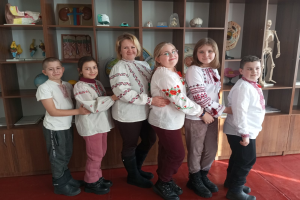 Gruppe von Kindern in traditioneller ukrainischer Kleidung vor einem Bücherregal mit Spielzeug, einem Skelett und anderen Gegenständen