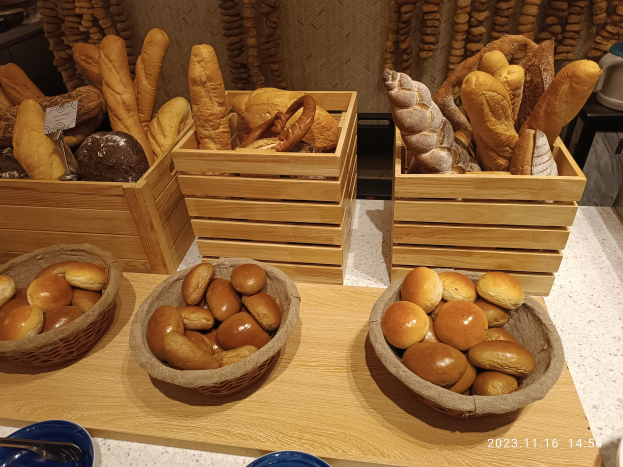 Ein Holztisch zeigt Körbe mit verschiedenen Brotsorten, Teller und Löffel, mit einer gewässerten Wand im Hintergrund, die auf eine Pariser Bäckerei hindeutet.