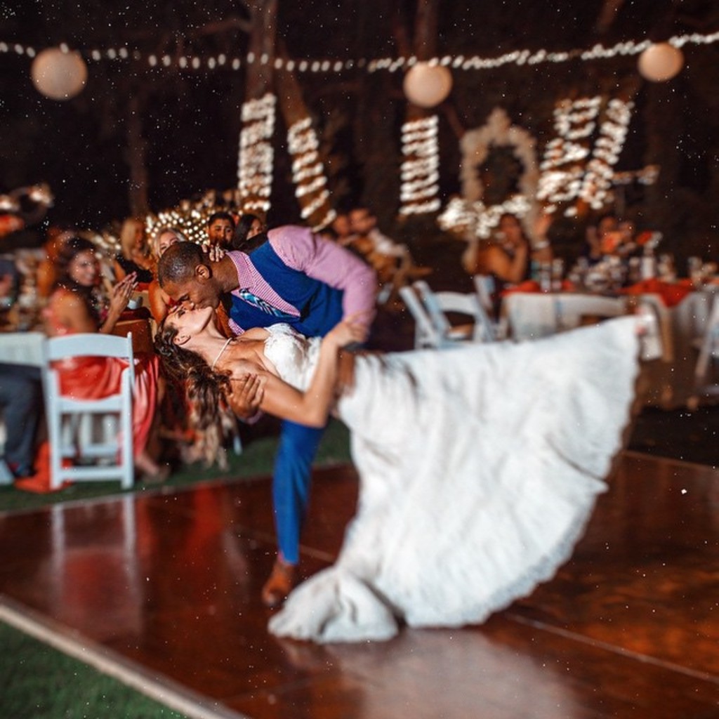 Eine Person küsst eine Frau in einer Outdoor-Szene mit Menschen an Tischen, Dekokugeln, Bäumen und Lichtern im Hintergrund.