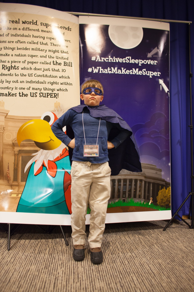 Ein junger Junge in einem Superheldenkostüm steht vor einem Banner mit Text und einem Vogelbild, mit einem Vorhanghintergrund und Schuhen.