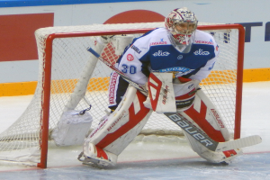 Ein Eishockeytorwart in voller Ausrüstung steht in seinem Netz auf dem Eis, mit einer textbedeckten Wand im Hintergrund.