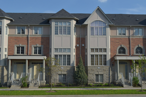 Eine Reihe von Stadthäusern mit Fenstern, Türen, Säulen, Geländern, Treppen, Pflanzen, Bäumen, Gras, einer Straße im Vordergrund und dem Himmel im Hintergrund.