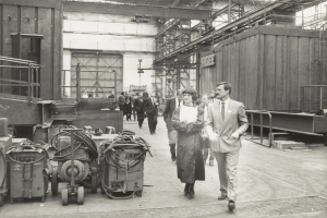 Eine Gruppe von Menschen in Anzügen und Schuhen, die durch eine Fabrik gehen, mit Maschinen auf der linken Seite und einer Wand im Hintergrund.