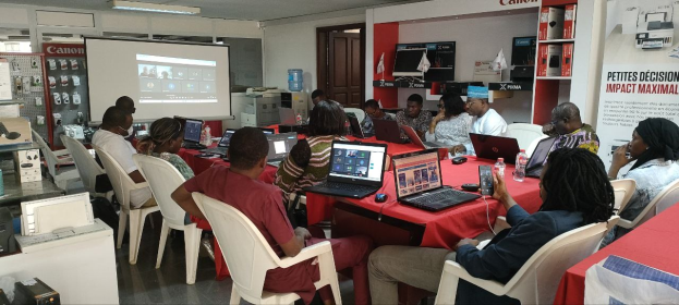 Gruppe von Menschen um einen Tisch mit Laptops und anderen Gegenständen während eines digitalen Marketing-Workshops, mit einem Banner, einem Display und einer Tür im Hintergrund.