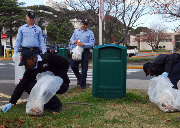 Drei Polizeibeamte in Uniform sammeln Müll an einer Straßenrand, mit Bäumen, Gebäuden, Fahrzeugen und einem Mülleimer im Hintergrund.