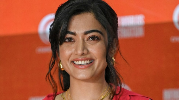 Frau in einem roten Kleid mit einem eleganten Dutt und dezentem Make-up, die strahlend in die Kamera lächelt, vor einem gelben Banner mit der Aufschrift "Sonam Kapoors beste Schönheits Looks vom Cannes Film Festival."