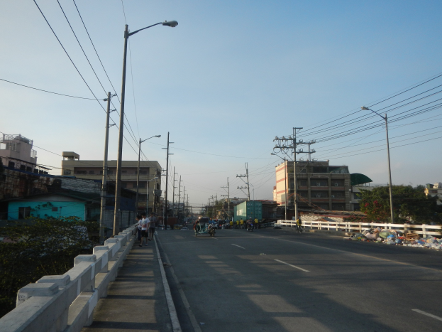 Stadtstraße mit Strommasten, Straßenlaternen, Kabeln, Fahrzeugen, Fußgängern, Geländern, Grünflächen, Gebäuden und herumliegendem Müll gegen den Himmel.