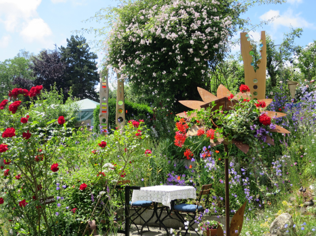 Ein Garten mit bunten Blumen und Pflanzen, der einen Tisch mit einer Decke und Stühlen zeigt, vor einem Wald und einem bewölkten Himmel auf der Chelsea Flower Show.