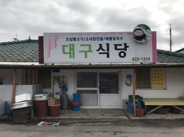 Kleines Gebäude mit einer Tafel, die "Koreanisches Restaurant" lautet, einer Tür, Fenstern, einem Dach und einem bewölkten Himmel, umgeben von Behältern, Eimern, einem Tisch, Pfählen und einem weiteren Haus im Hintergrund.