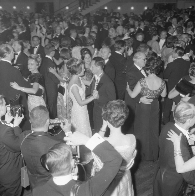 Ein Schwarz-Weiß-Foto von einer Gruppe von Menschen, die auf einer Tanzfläche tanzen, einige halten Kameras, mit Treppen und Lichtern im Hintergrund.