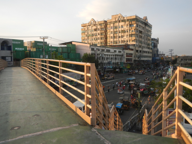 Ein Fußgängerbrücke mit Geländern überspannt eine vielbefahrene Straße voller Fahrzeuge und Fußgänger, mit Gebäuden, Bäumen, Strommasten und einem bewölkten Himmel im Hintergrund.