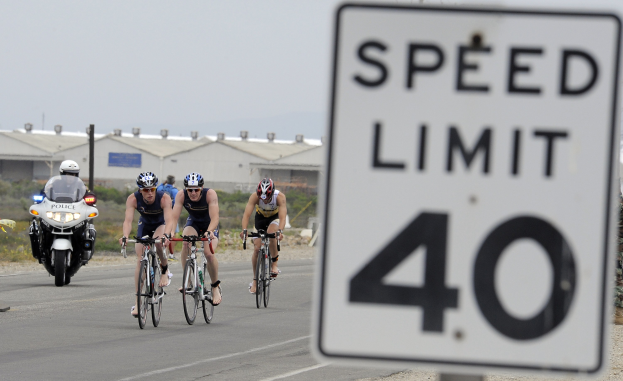 Gruppe von Radfahrern mit Helmen, die eine Straße mit einem Geschwindigkeitsbegrenzungsschild entlangfahren, mit Gebäuden und Bäumen im Hintergrund unter einem klaren blauen Himmel.